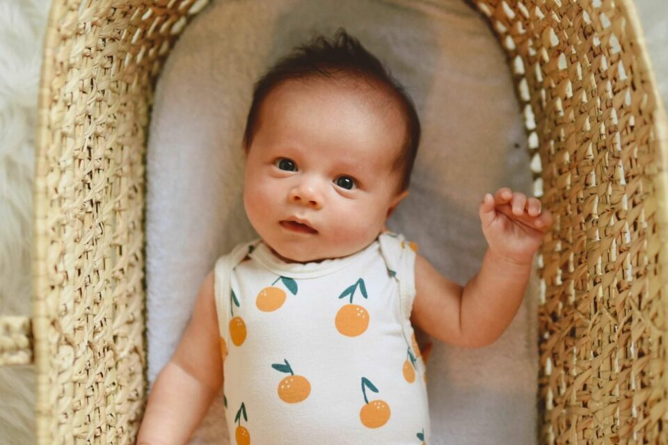 View from above of a baby wearing a baby grow decorated with oranges, lying on their back in a Moses basket.