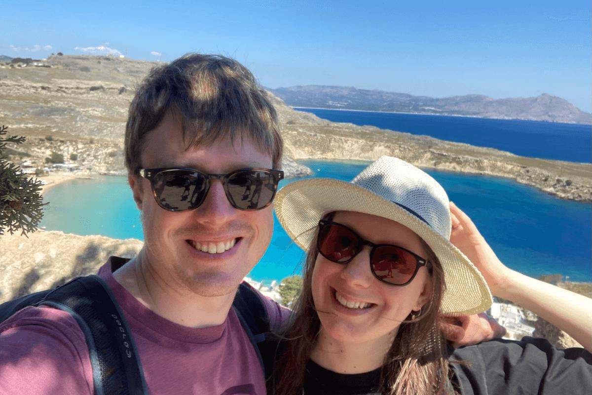 Laura and Matt are taking a selfie. They are on holiday with a strikingly blue sea behind them, wearing sunglasses and Laura wears a sun hat.