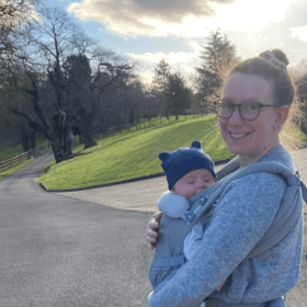 Laura and Sam are pictured outdoors. Laura is smiling with her hair in a bun, and Sam is strapped to her front, wearing a hat with teddy ears.