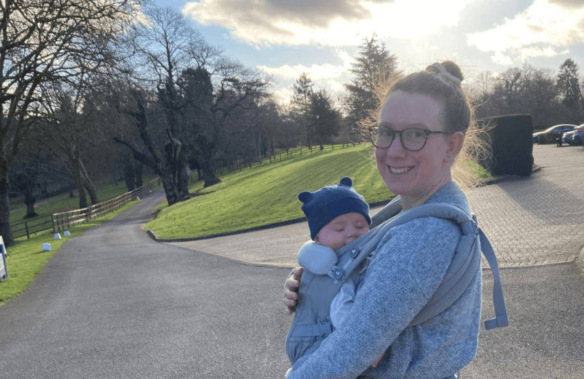 Laura and Sam are pictured outdoors. Laura is smiling with her hair in a bun, and Sam is strapped to her front, wearing a hat with teddy ears.
