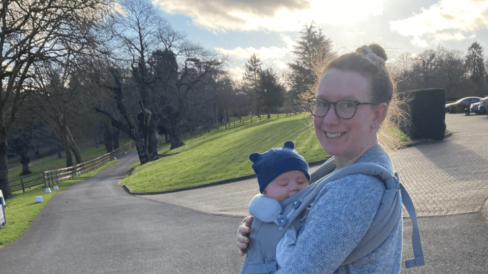 Laura and Sam are pictured outdoors. Laura is smiling with her hair in a bun, and Sam is strapped to her front, wearing a hat with teddy ears.
