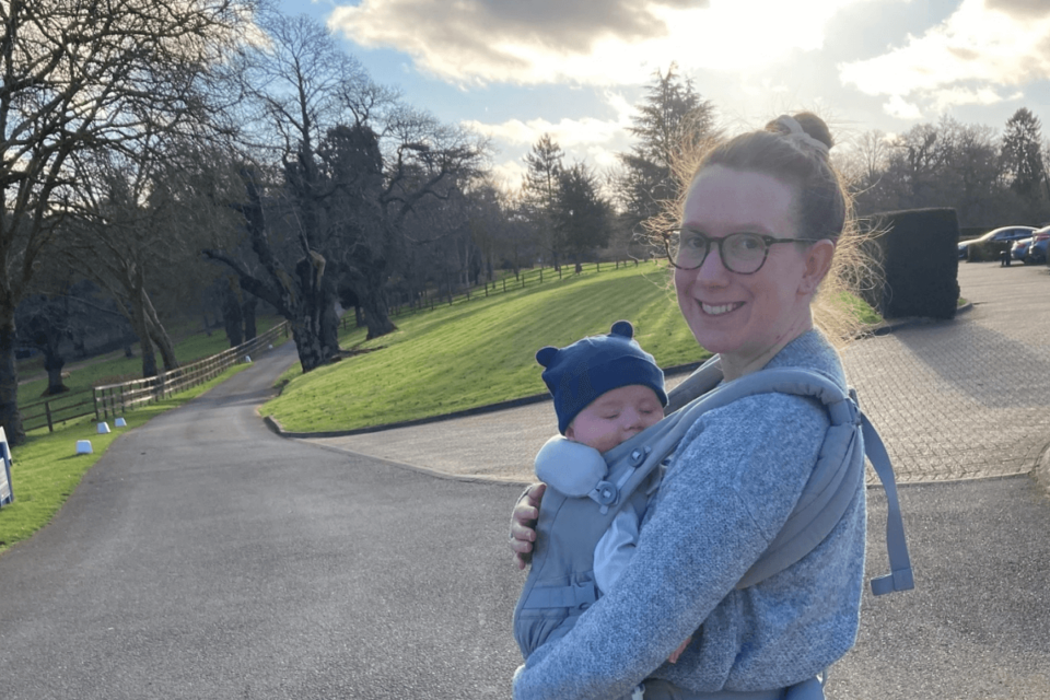 Laura and Sam are pictured outdoors. Laura is smiling with her hair in a bun, and Sam is strapped to her front, wearing a hat with teddy ears.