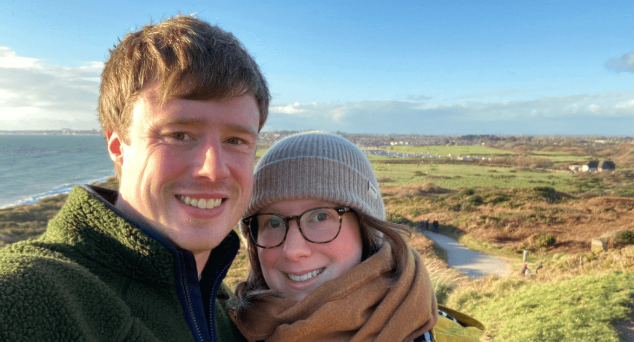 Matt and Laura are taking a selfie in front of green grassy moors. They are dressed warmly and both smiling.
