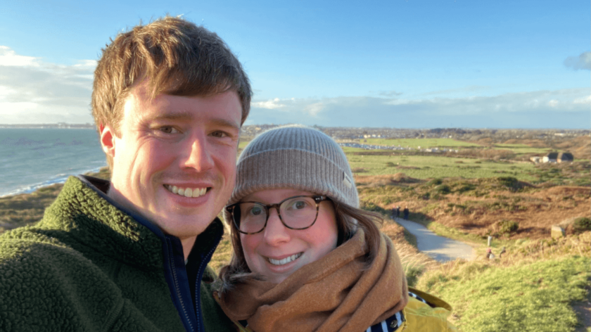 Matt and Laura are taking a selfie in front of green grassy moors. They are dressed warmly and both smiling.