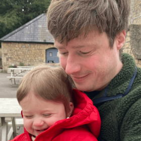 Matt and baby Sam are sat on a bench. Sam is grinning cheekily and Matt is holding him, smiling and looking down at baby Sam's hands.