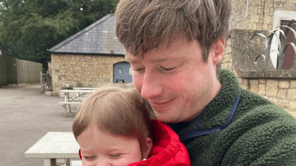 Matt and baby Sam are sat on a bench. Sam is grinning cheekily and Matt is holding him, smiling and looking down at baby Sam's hands.