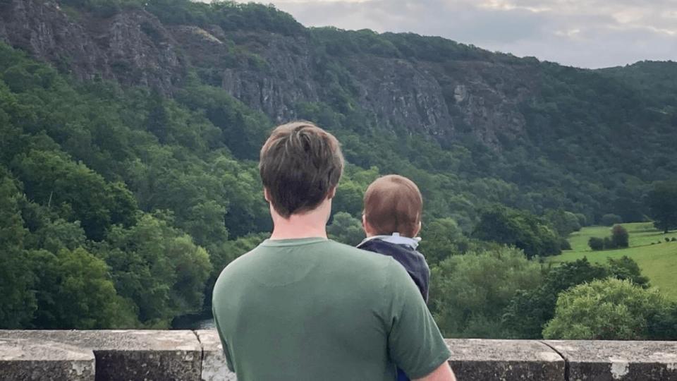 Baby Sam is being held by Matt's right arm. They are both looking out over a bridge at a beautiful, green view. Their backs are to the camera.