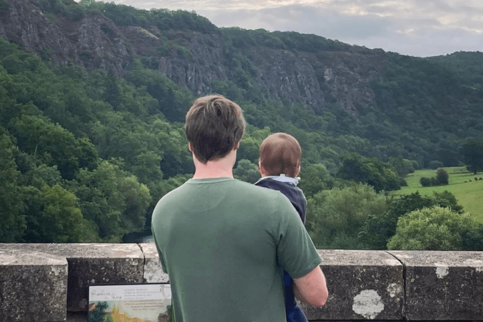 Baby Sam is being held by Matt's right arm. They are both looking out over a bridge at a beautiful, green view. Their backs are to the camera.