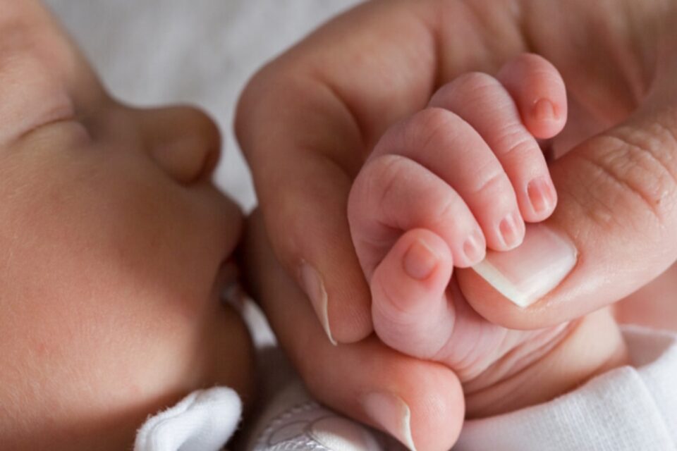 A baby is sleeping and their parent is holding their hand. The baby is gripping their parent's thumb,