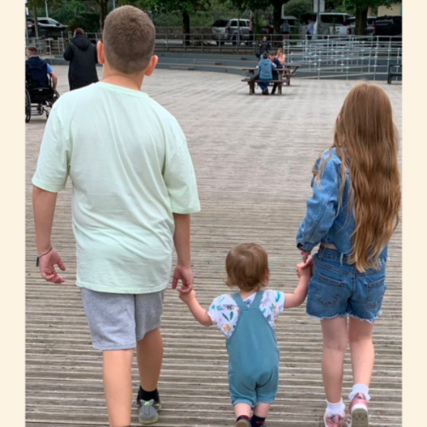 Bailey Poppy and Jack are walking with their backs to the camera. The eldest are holding the hands of the little one, Jack's, hand in the middle. They are dressed in summer clothes.