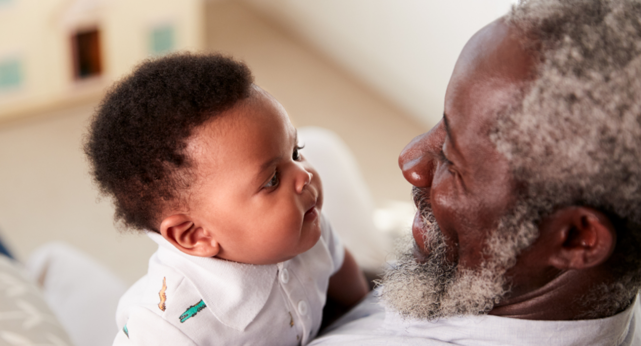 A baby is looking at his grandparent and their faces are close together. The granddad has dark skin and hints of grey in their hair and bears, and the baby is wearing a baby grow.