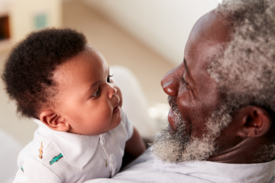 A baby is looking at his grandparent and their faces are close together. The granddad has dark skin and hints of grey in their hair and bears, and the baby is wearing a baby grow.