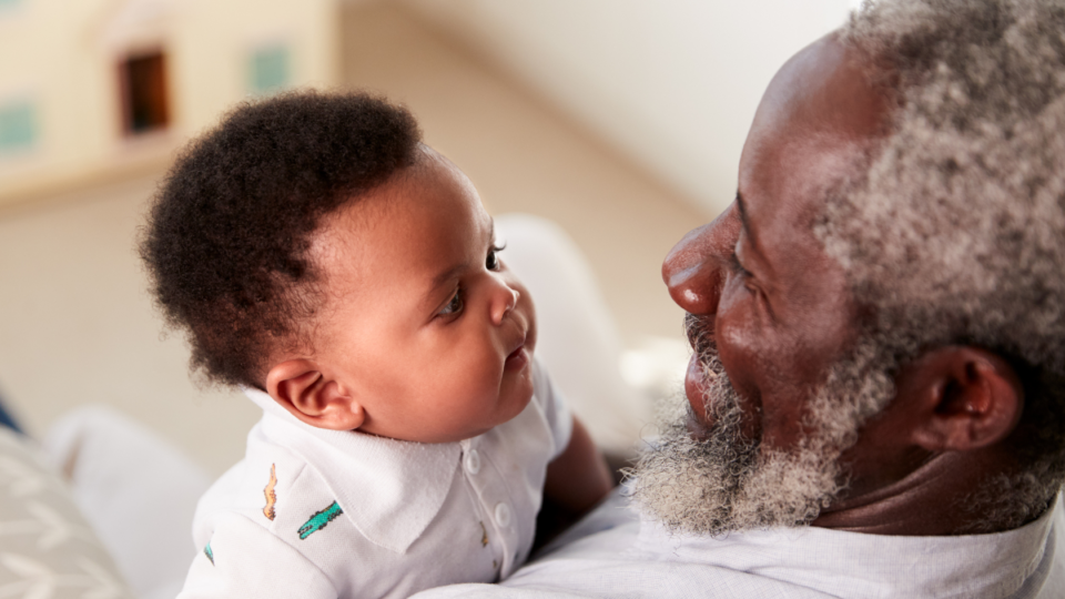 A baby is looking at his grandparent and their faces are close together. The granddad has dark skin and hints of grey in their hair and bears, and the baby is wearing a baby grow.