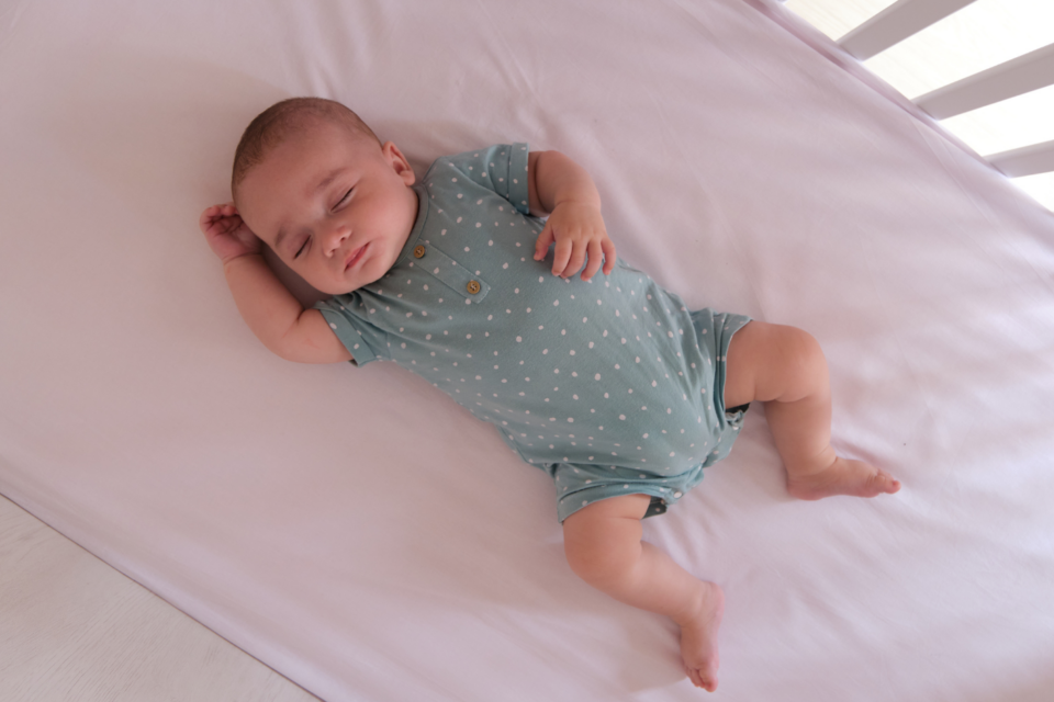 Baby sleeping in a cot on their back. They have a light sleep suit on and the light is coming through the bars of the cot. They have light skin.