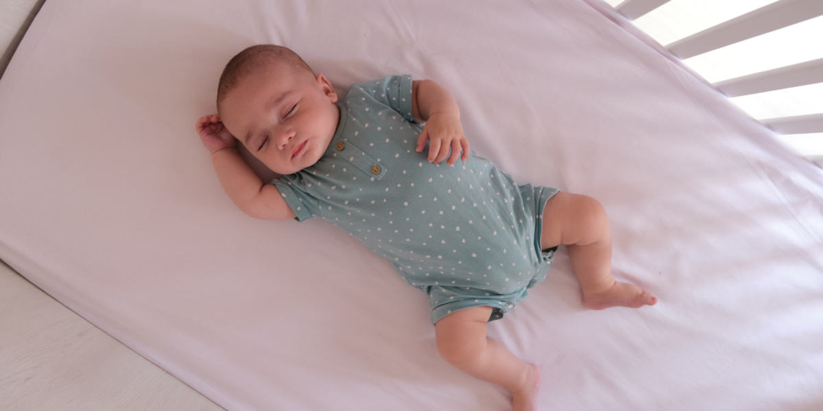 Baby sleeping in a cot on their back. They have a light sleep suit on and the light is coming through the bars of the cot. They have light skin.