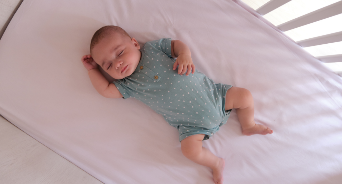 Baby sleeping in a cot on their back. They have a light sleep suit on and the light is coming through the bars of the cot. They have light skin.