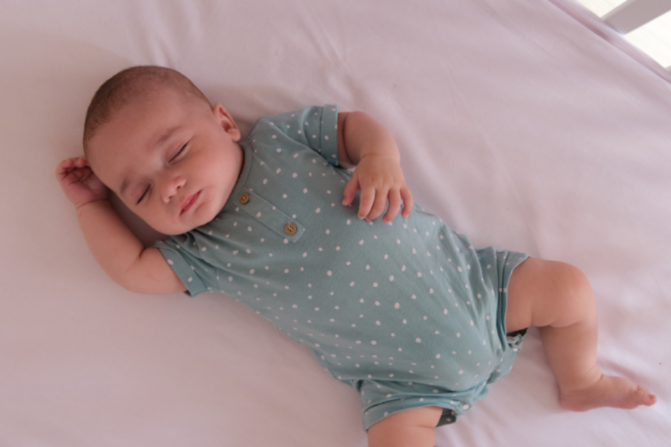 Baby sleeping in a cot on their back. They have a light sleep suit on and the light is coming through the bars of the cot. They have light skin.