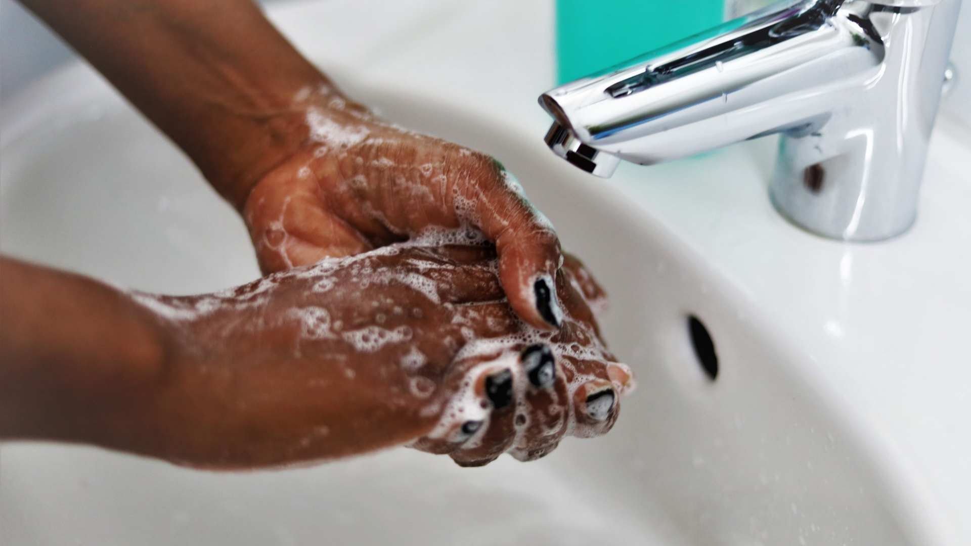 A close up of a woman washing her hands. She has soap suds on her hands and black nail polish, and the tap is off.