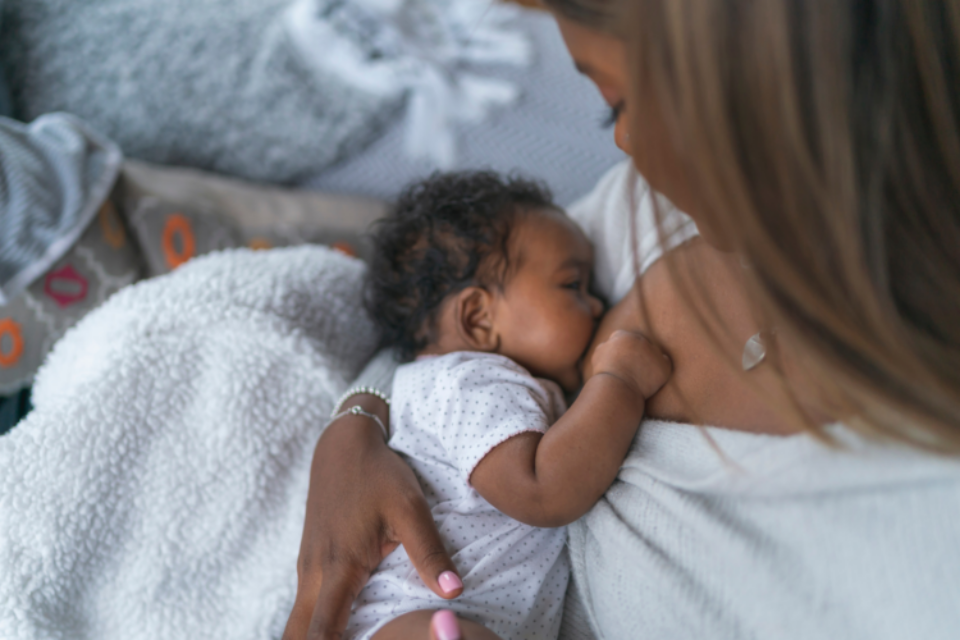 A baby is being breastfed by their mum. They look very young, have curly dark hair and dark skin. Their mum is looking down at them.