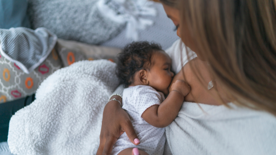 A baby is being breastfed by their mum. They look very young, have curly dark hair and dark skin. Their mum is looking down at them.