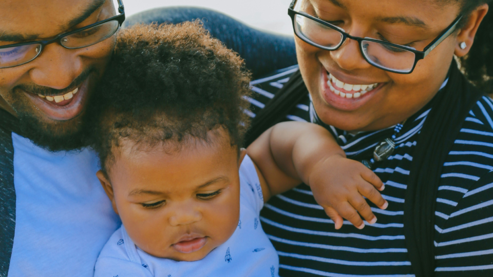 A family are pictured together close-up. They have dark skin and both mum and dad are smiling down at their baby. Their baby has the same skin and curly, dark brown hair and is looking down at something.