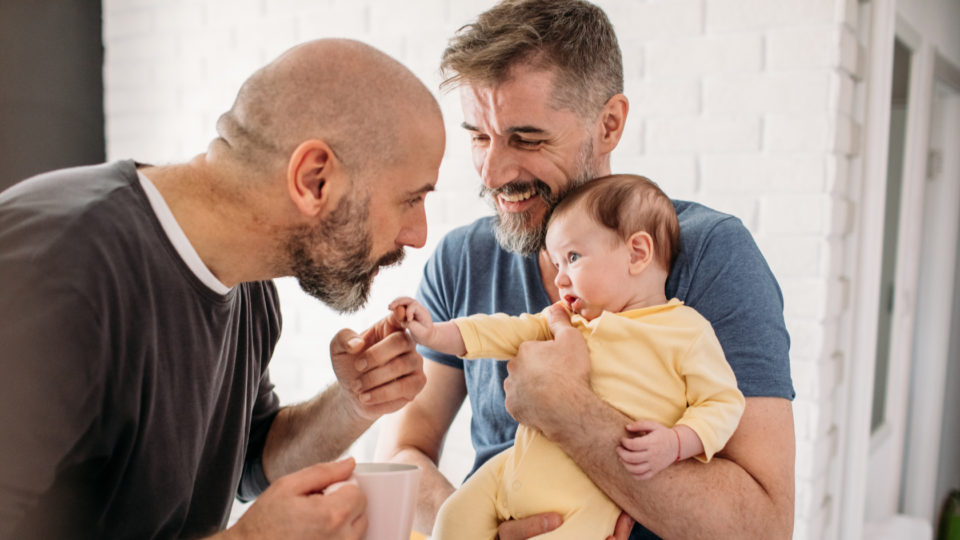 One man is holding a baby with the baby's head near his chin. The other man is holding the baby's hand in his, and putting his mouth to the baby's hand. Both men are smiling.