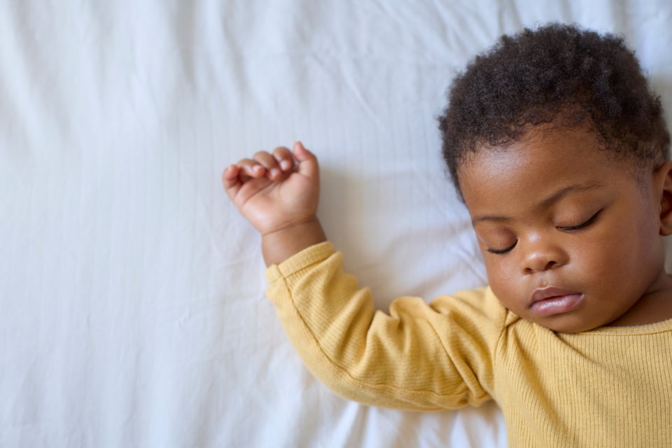 A baby is sleeping on their back. They have a yellow sleepsuit on and they have dark skin with curly hair.