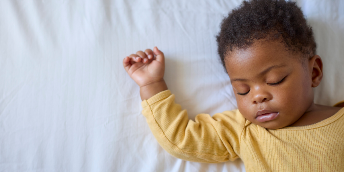 A baby is sleeping on their back. They have a yellow sleepsuit on and they have dark skin with curly hair.