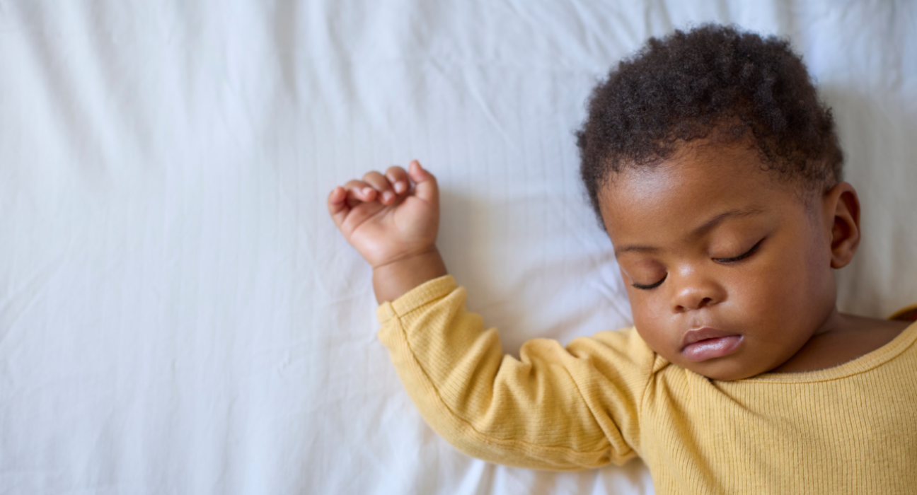 A baby is sleeping on their back. They have a yellow sleepsuit on and they have dark skin with curly hair.