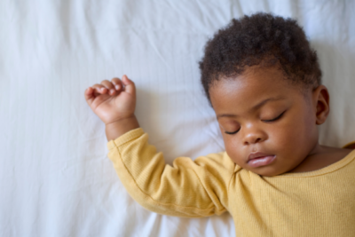 A baby is sleeping on their back. They have a yellow sleepsuit on and they have dark skin with curly hair.
