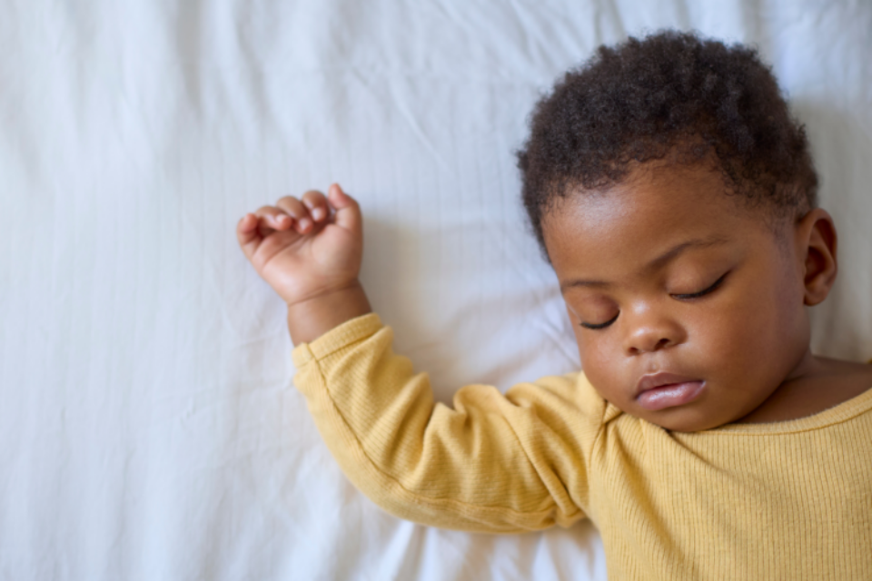 A baby is sleeping on their back. They have a yellow sleepsuit on and they have dark skin with curly hair.