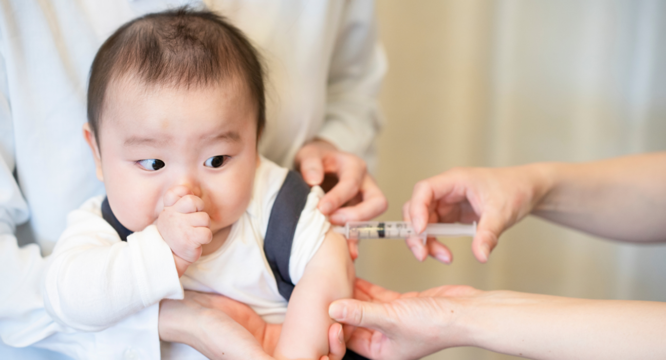A baby is getting one of their early vaccinations. They are sucking their thumb as the nurse inserts a needle into their arm, administering the vaccine. They are being held by a parent.