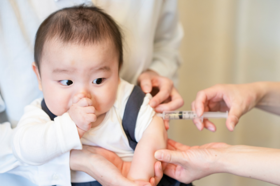 A baby is getting one of their early vaccinations. They are sucking their thumb as the nurse inserts a needle into their arm, administering the vaccine. They are being held by a parent.