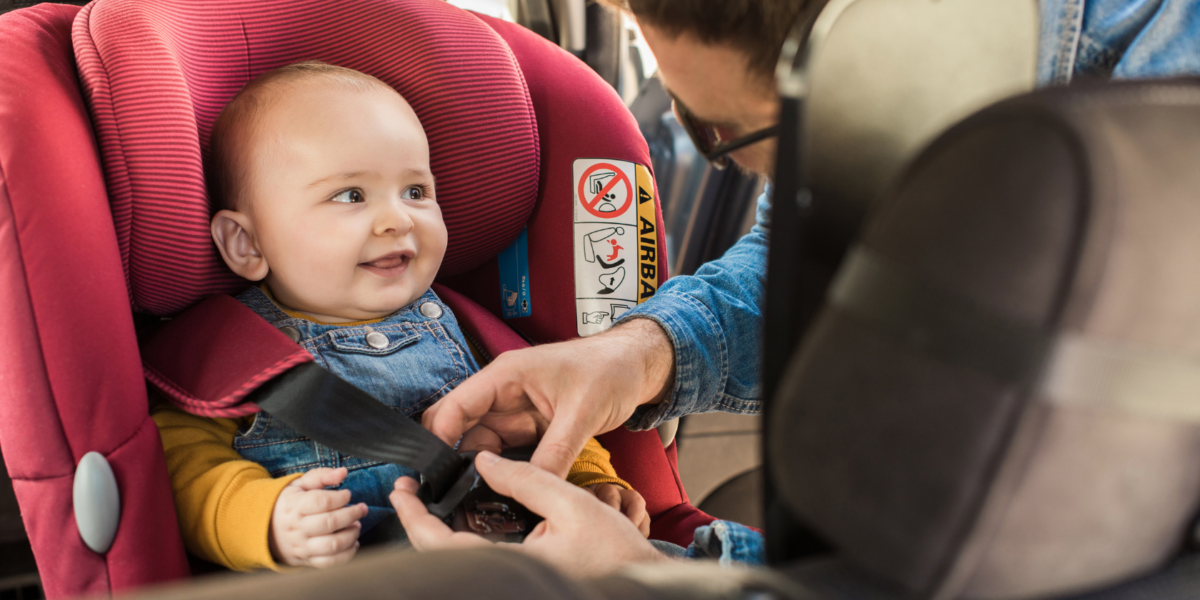 A baby is being fastened into a car seat by their parent. They have light skin, a small amount of light hair and are looking to the side, smiling at their parent.