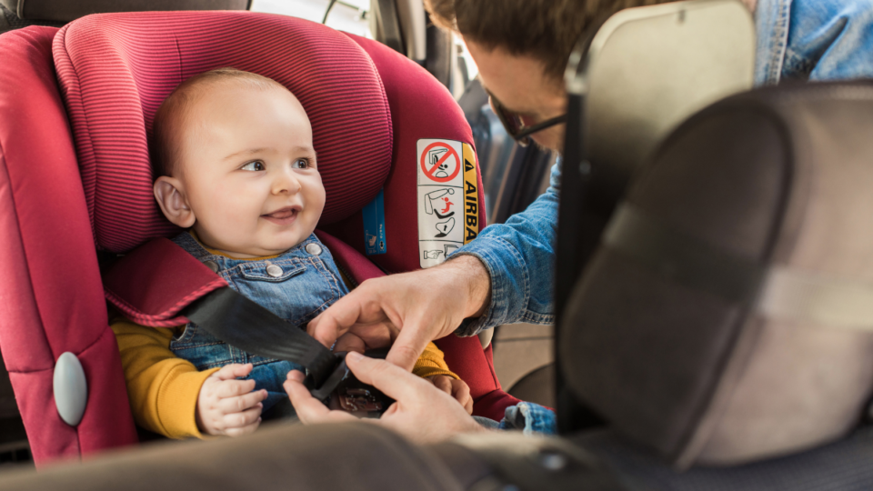 A baby is being fastened into a car seat by their parent. They have light skin, a small amount of light hair and are looking to the side, smiling at their parent.