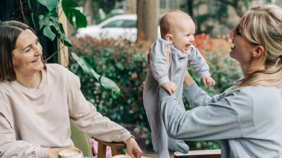 Two women are sat at a cafe and one is holding the baby up in their arms. They are both smiling, but the baby has the biggest and widest smile of them all. They have light skin and the baby is wearing a baby grow.