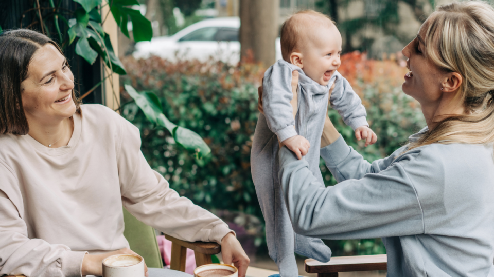 Two women are sat at a cafe and one is holding the baby up in their arms. They are both smiling, but the baby has the biggest and widest smile of them all. They have light skin and the baby is wearing a baby grow.