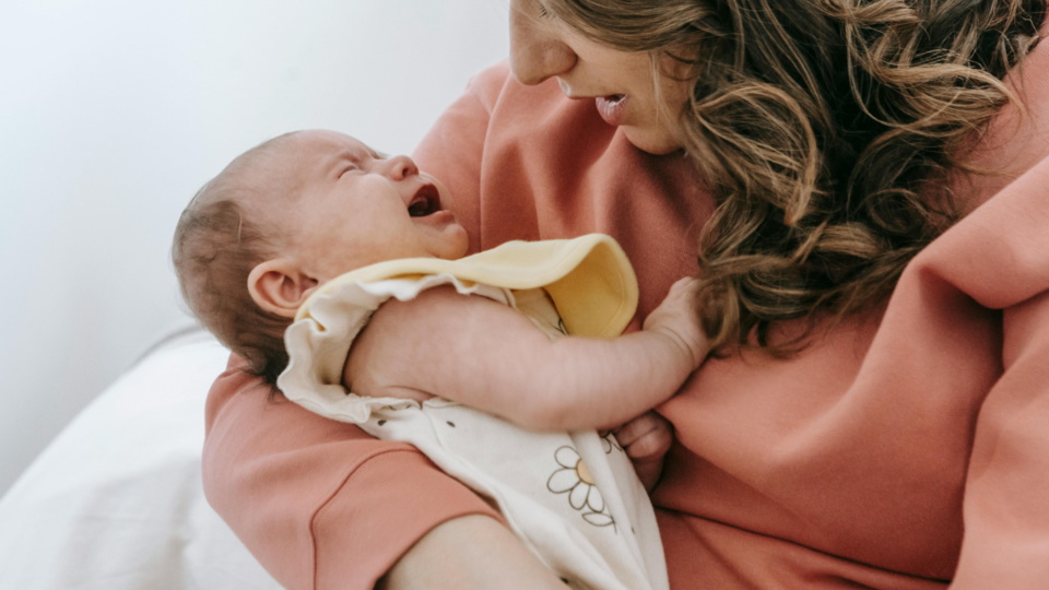 A baby is crying in their mum's arms. They have light skin and wear a floral baby grow. You can only see part of mum's face but can make out her brunette hair.