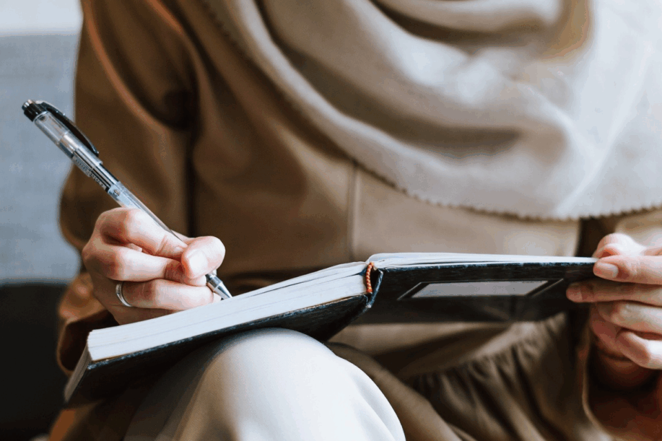 Woman wearing a hijab, close up of her hand writing in a notebook using a pen.
