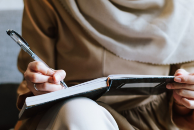 Woman wearing a hijab, close up of her hand writing in a notebook using a pen.