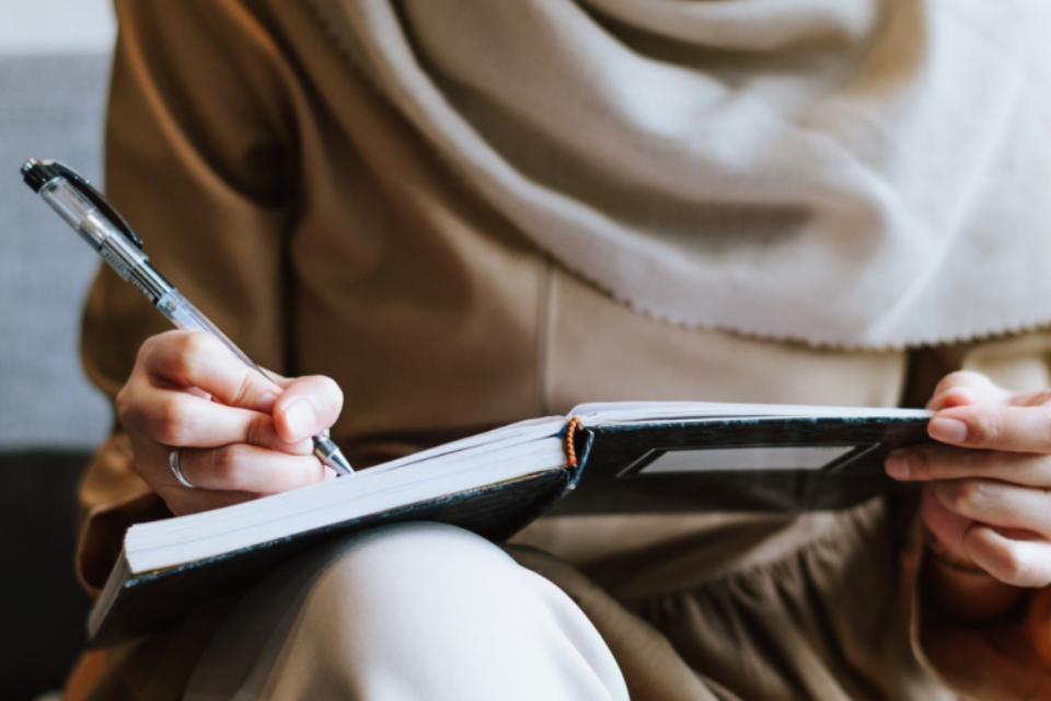 Woman wearing a hijab, close up of her hand writing in a notebook using a pen.