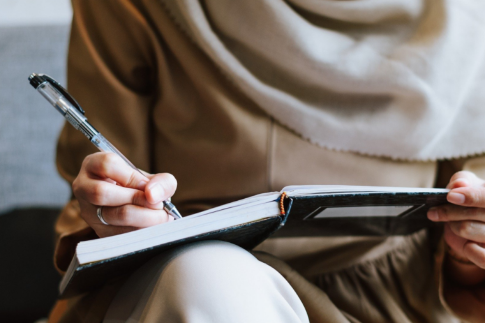 Woman wearing a hijab, close up of her hand writing in a notebook using a pen.