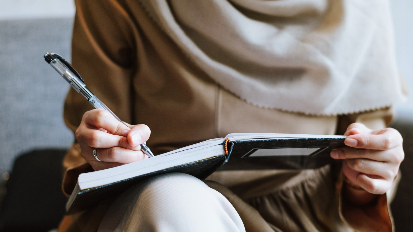 Woman wearing a hijab, close up of her hand writing in a notebook using a pen.