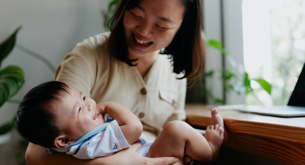 A mum is holding her baby in her arms. The baby is laughing and giggling with their hand in their mouth, and the mum is looking down at the baby, laughing too,