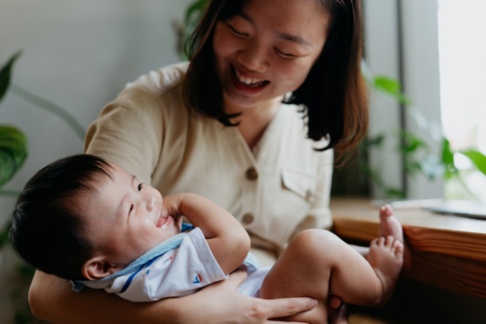 A mum is holding her baby in her arms. The baby is laughing and giggling with their hand in their mouth, and the mum is looking down at the baby, laughing too,