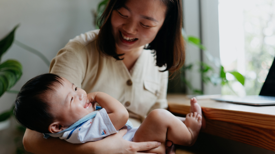 A mum is holding her baby in her arms. The baby is laughing and giggling with their hand in their mouth, and the mum is looking down at the baby, laughing too,