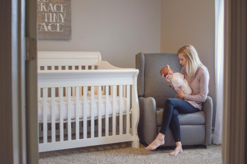 A young baby is in a swaddle next to their empty cot (safer sleep). The baby is being held by their mother who is settling the baby to sleep on their armchair.