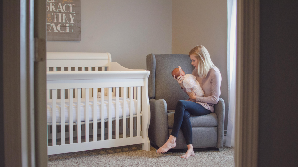 A young baby is in a swaddle next to their empty cot (safer sleep). The baby is being held by their mother who is settling the baby to sleep on their armchair.