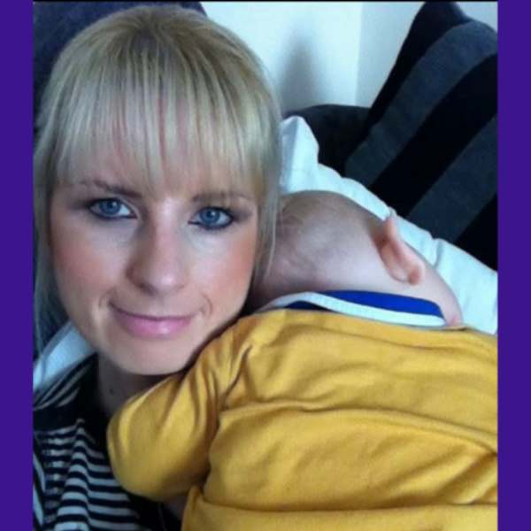 Karen has a blonde fringe and a baby is draped on her shoulder, peacefully sleeping. She is grinning at the camera and has striking eye makeup.