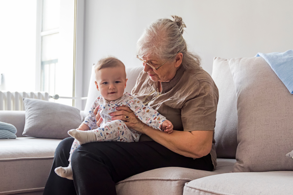 An older woman is sat with a young baby on her lap. She is looking down at the baby and the baby is smiling.
